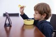 © Татьяна Волкова - Brown haired boy sit at table and with interest watch cartoon on tablet computer side view. Kid of kindergarten age in raised hand hold spoon to eat dry chocolate balls. Gadget addiction, habit