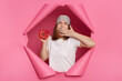 Horizontal shot of sleepy tired young woman wearing white T-shirt and sleeping mask breaking through paper hole of pink background, showing alarm clock, yawning, covering mouth with hand.