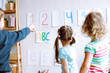 © Татьяна Волкова - Children stand with back and learn english letters, alphabet and numbers on cards on wall. Kids point at colored numbers on sheets of paper by finger. Reading and counting, study in daycare centre