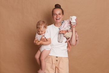 Indoor shot of smiling happy woman mother wearing white T- shirt standing with her little daughter in hands isolated over brown background, drinking coffee in the morning.