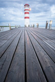 Fototapeta Zachód słońca - Red and white lighthouse on a cloudy morning with wooden jetty leading to it, vertical, Austria