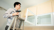 © zephyr_p - Asian male furniture assembler using electric drill screwdriver on cabinet hinge of glass panel. Interior construction worker man standing on ladder using screwdriving tools for furniture installation