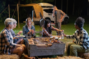 Group of family camper van sitting by fire grilling sausages and drinking dancing having dinner