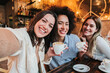 © Jose Calsina - Group of three happy young women taking a selfie portrait on a coffee shop. Multi ethnic friends taking a photo smiling and looking at camera. High quality photo