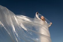 Portrait Of Blonde Girl In White Dress Waving Plastic Sheet Under Sunny Blue Sky