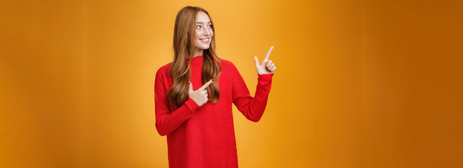 Wall Mural - Interested gentle and tender young 20s ginger woman in red warm sweater pointing, looking at upper left corner with broad smile, enjoying nice happy scene over orange background