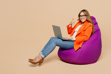 Full body young employee IT business woman corporate lawyer in classic formal orange suit glasses work office sit in bag chair use laptop pc computer show thumb up isolated on plain beige background