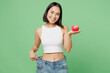 Leinwandbild Motiv Young woman wears white clothes show loose pants on waist after weightloss hold red apple isolated on plain pastel light green background. Proper nutrition healthy fast food unhealthy choice concept.