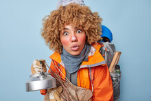 Photo Of Shocked Curly Haired Woman Carries Kettle And Wood Applies Protective Facial Cream On Frozen Skin Poses With Rucksack Has Camping Trip Isolated Over Blue Background. Hiking And Backpacking