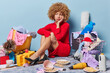 Unhappy stressed lady wears red dress and black high heeled shoes sits on floor against different items around upset because of chaos in house looks sadly away isolated over blue background.