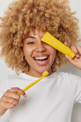 Wall Mural - Stomatology and health care concept. Positive curly haired woman covers eye with toothpaste holds yellow toothbrush takes care of her dentals tries to prevent caries laughs joyfully. Toothcare routine