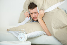 Man In Bed By Alarm Clock Holding Pillow Over Head