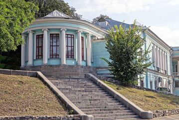 Sticker - facade of a blue old building with white stone columns and red windows on a city street among green vegetation near a gray staircase