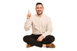 © Asier - Adult latin man sitting on the floor cut out isolated showing number one with finger.