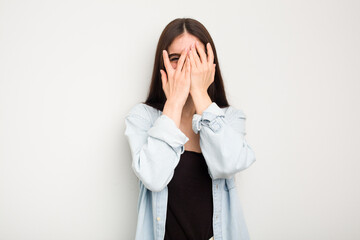 Wall Mural - Young caucasian woman isolated on white background blink at the camera through fingers, embarrassed covering face.