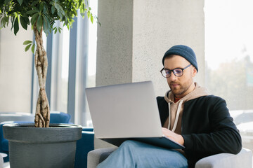 Young handsome freelancer guy working with laptop in office space. A man in glasses and a hat is holding a laptop while sitting in a chair in the hall.