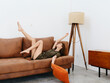 © SHOTPRIME STUDIO - Young woman lying on orange couch at home with her legs up and tossing pillow, happy relaxation at home, stylish interior, copy space