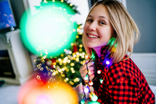 Smiling Girl Wearing Multi Colored Illuminated String Lights At Home