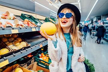 Stylish fashion laughing woman holding fresh organic melon in the supermarket store during selecting fresh products. Veganuary month, Healthy eating diet, go vegan. Selective focus. Copy space.