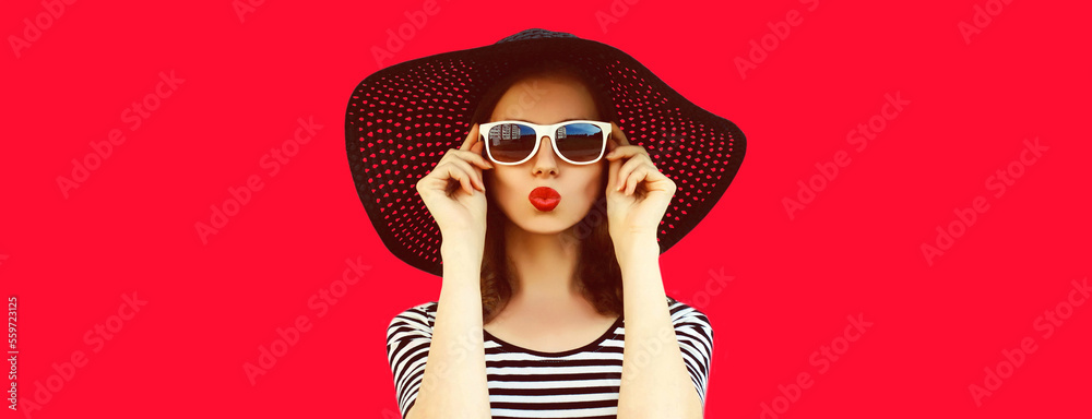 Portrait of beautiful young woman blowing her lips with red lipstick sending sweet air kiss wearing black summer straw hat on background