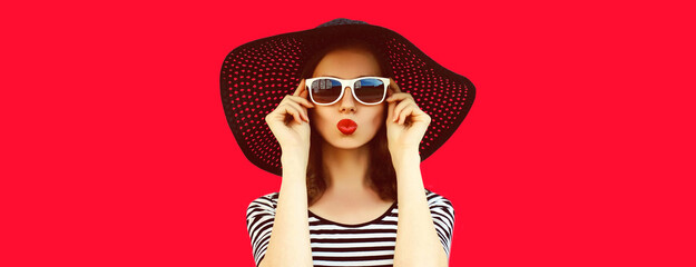 Portrait of beautiful young woman blowing her lips with red lipstick sending sweet air kiss wearing black summer straw hat on background