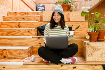 Wall Mural - Young asian woman drinking coffee and using laptop on stairs indoors