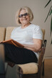 © Strelciuc - Creative senior female writer wearing eyeglasses sitting comfortably on sofa with copybook and pen, making notes, smiling at camera. Business people lifestyle.