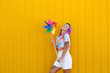 © VISTA by Westend61 - Smiling woman holding multi colored pinwheel toy in front of yellow wall
