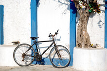 Bicycle On The Street In Greece Style With White Wall And Blue Frames And Old Tree 