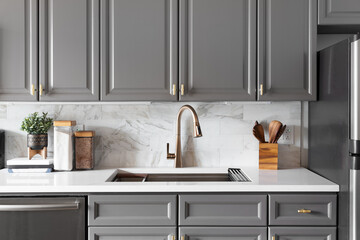 A kitchen sink detail shot with grey cabinets, a white marble countertop and backsplash, and decorations.