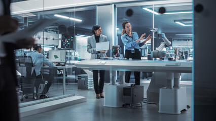 Poster - Two Female Working in High Tech Facility. Robotics Engineer and Machinery Operator Collaborate on a Robot Arm Project, Using a Laptop Computer to Operate the Robot Arm to Move Around a Microchip.