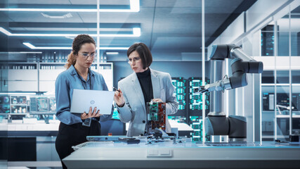 Leinwandbilder - Two Female Working in High Tech Facility. Robotics Engineer and Machinery Operator Collaborate on a Robot Arm Project, Using a Laptop Computer to Operate the Robot Arm in Technology Startup Lab.