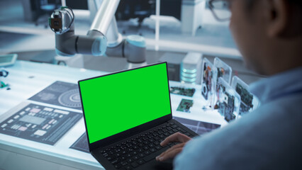 Fototapete - Over the Shoulder Shot of a Robotics Engineer Using a Laptop Computer with Green Screen Mock Up Display Screen. Robotic Arm Holding a Microchip. Automation Startup Research and Development Office.
