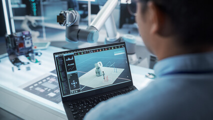 Over the Shoulder Shot of a Robotics Engineer Using a Laptop Computer with a Software Operating the Robotic Arm. Robot Hand Holding a Microchip. Automation Startup Research and Development Office.