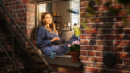 Wall Mural - Female Manager Writing her Work Emails Using a Laptop While Sitting on her Windowsill. Young Beautiful Woman Enjoying Calmness and Comfort Thanks to the Possibility to do Remote Job