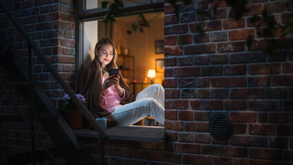 Wall Mural - Happy Beautiful White Woman Using her Smartphone While Sitting on her Windowsill During Evening. She's Browsing the Internet and Checking Videos on Social Networks and Having Fun