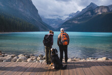 Young Couple With Backpacks And A Dog. Tourists And Travelers Exploring Lake Louise In Banff, Canada.