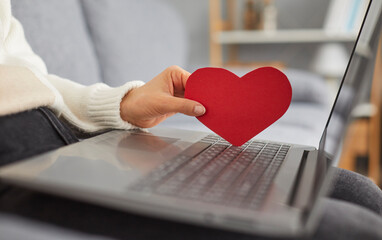 Young woman is looking for love online and using a dating website. Cropped shot of an unrecognisable lady with a laptop computer on her lap holding a red paper heart. Online dating concept