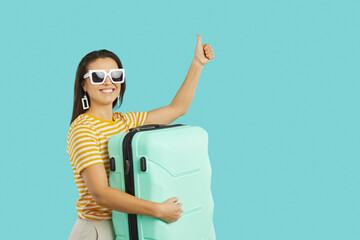 Wall Mural - Happy tourist going on holiday trip and giving thumbs up. Cheerful beautiful woman in sun glasses isolated on turquoise background holding suitcase, doing like gesture and smiling. Travelling concept