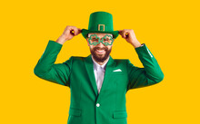 Cheerful Man Going To Party On St Patrick's Day. Happy Smiling Joyful Positive Young Guy Wearing Green Suit, Funny Shamrock Glasses And Leprechaun Top Hat Standing Isolated On Yellow Color Background