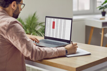 Concentrated Male Financier Using Laptop Working In Office With Data Entered In Spreadsheet. Man Fills Out Paper Documents And Enters Data Into Electronic Files Marked In Red On Laptop Screen.