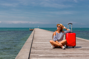 A lady is sitting on a pier against the background of the sea with a red suitcase for luggage, shorts, sunglasses, travel 2023, summer mood, open borders