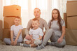 © lenblr - Family with excited and amused expressions sits on floor among unpacked boxes and enjoys moving into new apartment. Brothers and parents smile
