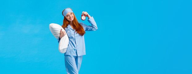 Wall Mural - Time to bed. Vertical full-length cheerful lovely, gentle redhead european woman with long red hair, wearing nightwear and sleep mask, holding pillow and red alarm clock, sweet dreams