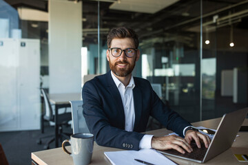 Portrait of confident middle aged businessman sitting at worktable in office, typing on keyboard, looking at camera