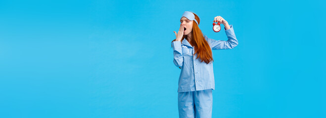 Wall Mural - Its so late time to bed. Tired and sleepy cute redhead female in nightwear and sleep mask, holding red alarm clock, yawning turn away with closed eyes, getting ready to sleep, blue background