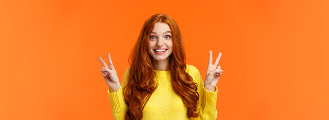 Wall Mural - Waist-up portrait lovely and cheerful pretty redhead woman showing peace gestures, smiling camera, travel abroad during winter holidays posing near sightseeing, orange background