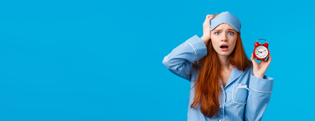 Wall Mural - Anxious, panicking young redhead girl missed interview, overslept, holding red alarm clock and touching head frustrated with concerned face, wearing nightwear and sleep mask, blue background