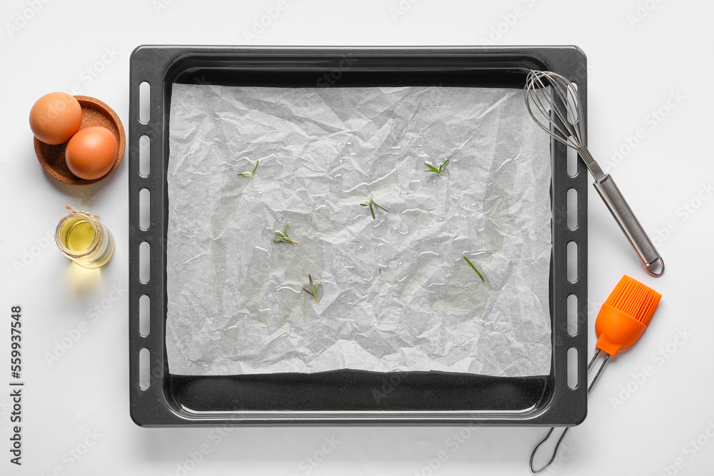 Baking tray with paper, eggs, oil, whisk and brush on white background