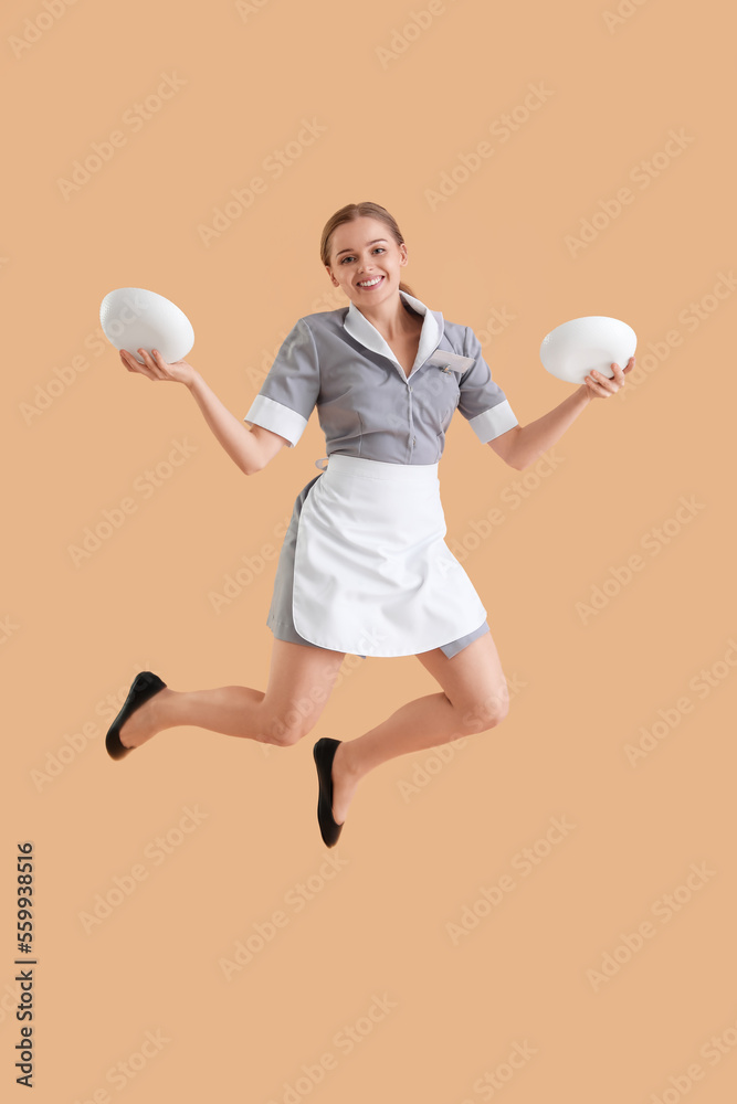 Young waitress with plates jumping on beige background